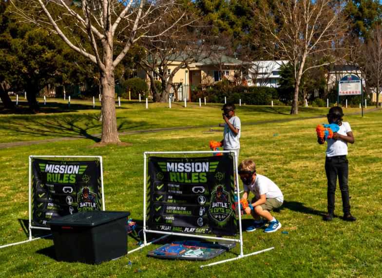 Kids playing Nerf battles behind Battle Ops Mission Rules barriers at a park party